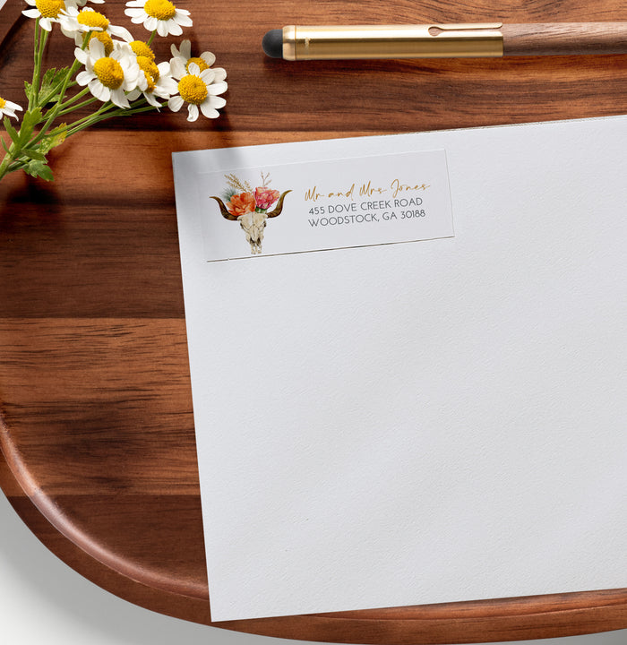 White envelope with floral design and text on a wooden surface with flowers and a pen.